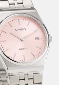 Montre en acier inoxydable avec un cadran rose, index argentés, fenêtre de date à 3 heures, et un bracelet en maillons métalliques. Étanchéité : 50M.
