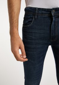 Tummansininen farkkuhousut, joissa on slim-fit -leikkaus. Housuissa on viisi taskua, hienovaraista kulumista ja kontrastiväriä keltaiset ompeleet saumoissa.