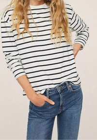 Camisa blanca de manga larga con rayas horizontales negras, combinada con jeans de mezclilla azules. La camisa tiene un corte holgado y un cuello redondo.
