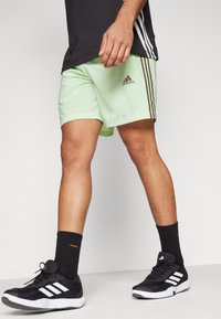 Ljust gröna sportshorts med en slät textur, med bruna sidostripes och en Adidas-logga. Kombineras med svarta träningsskor och strumpor.