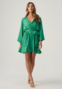 Grünes Seidenkleid mit tiefem V-Ausschnitt, Rüschensaum, langen Ärmeln und Taillenschnürung, das einen ausgestellten Saum und eine glatte Textur aufweist.