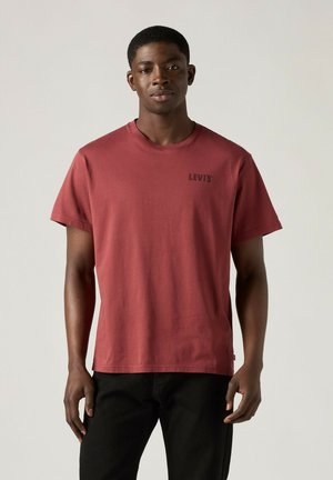 Hombre de pie que lleva una camiseta Levi’s color rojo ladrillo y pantalones negros contra un fondo claro liso.