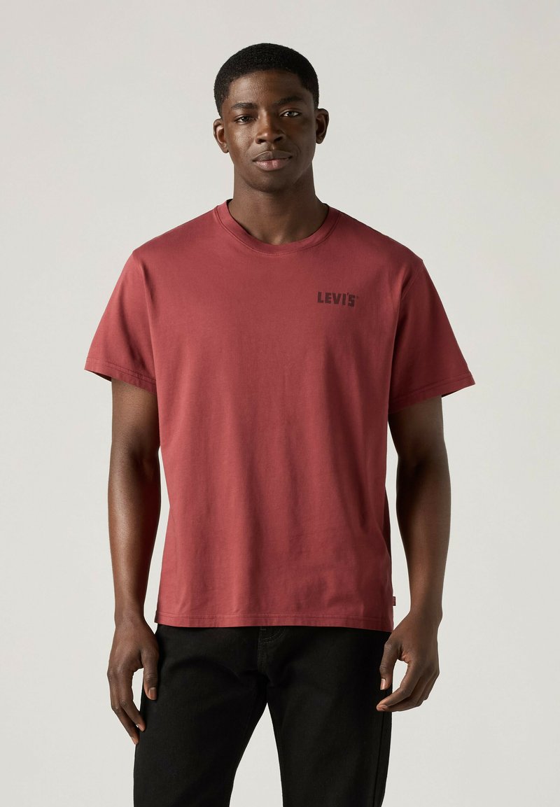 Mann steht, trägt ein ziegelrotes Levi’s T-Shirt und schwarze Hose vor einem einfarbig hellen Hintergrund.