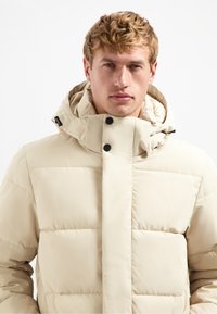 Beige pufferjack met een capuchon, voorzien van een drukknopen-sluiting, een gewatteerd ontwerp en een gladde textuur. Opvallende zwarte hardwareaccenten.