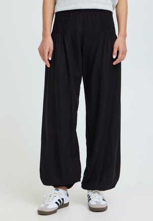 Kvinne iført løse svarte bukser med rynket midjebånd og hvite joggesko med svarte striper, stående mot en ensfarget bakgrunn.