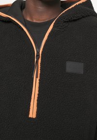 Pullover in pile nero con zip frontale, dotato di una cerniera arancione e rifiniture arancioni, tessuto testurizzato e un piccolo patch logo nero.