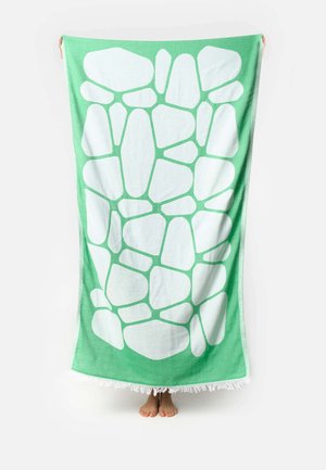 Groen-witte strandhanddoek met abstracte onregelmatige vormen, vastgehouden door een persoon, alleen zijn voeten zijn zichtbaar onder de handdoek.