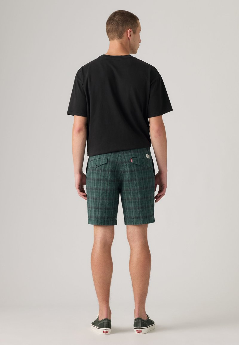 Uomo di spalle che indossa una t-shirt nera, pantaloncini verdi a quadri con tasche sul retro e scarpe slip-on verde scuro su uno sfondo semplice.