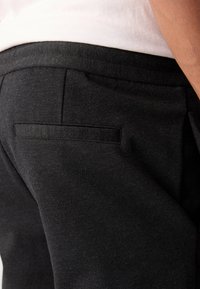 Des pantalons tissés noirs présentent un design élégant avec une poche arrière plate et un tissu texturé. La taille est finie avec un bord lisse.