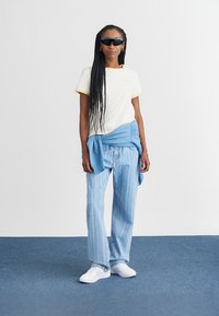 Weißes T-Shirt mit gelben Akzenten, hellblauer Pullover um die Taille gebunden, hell gestreifte Jeans und weiße Sneakers, dazu dunkle Sonnenbrille.