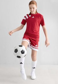 Rød atletisk skjorte med hvite detaljer og logo, røde shorts med hvite striper, hvite sokker, og en svart-hvit fotball på foten.