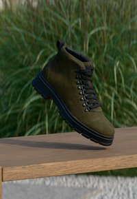 Olívovo zelená turistická obuv zo semišu s čiernymi šnúrkami a hrubou podrážkou, vznášajúca sa nad drevenou lavicou vonku s vysokou trávou v pozadí.