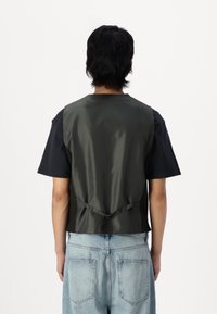 T-shirt nera a maniche corte sovrapposta a un gilet verde scuro in raso. Il gilet presenta un cordino di regolazione in vita e una texture liscia. Jean azzurri visibili.