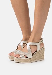 Sandali wedge in oro con fasce incrociate, tacco in juta intrecciata e chiusura con fibbia. Finitura metallica liscia e design open-toe.