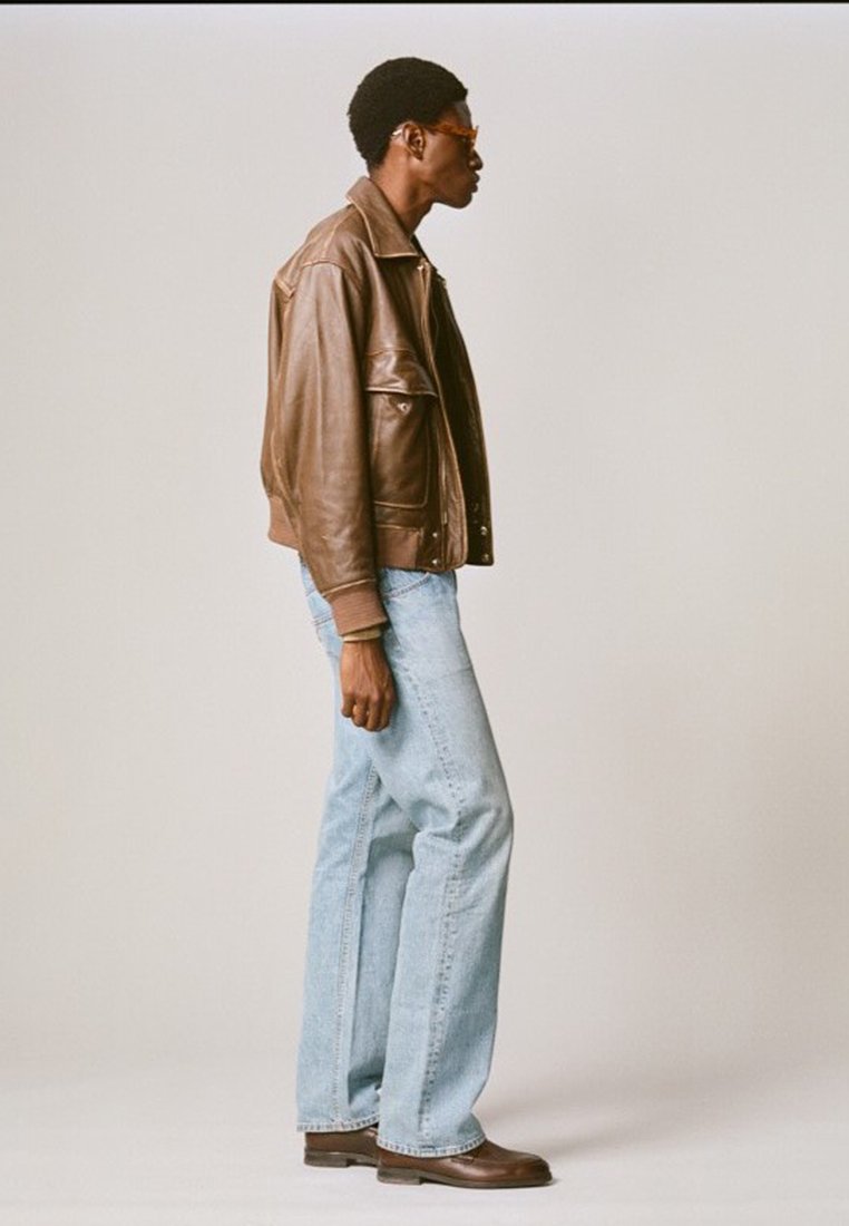 Giubbotto di pelle marrone, jeans di denim blu chiaro, stivali marroni e occhiali con lenti arancioni. Modello posizionato di lato contro uno sfondo neutro.