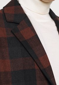 Hnědá a černá kostkovaná látka kabátu s jemnou texturou, s klopovým límcem a krémovým rolákem uvnitř.