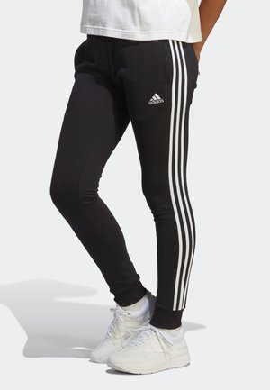 Personne portant un pantalon de survêtement Adidas noir avec trois bandes blanches sur les côtés et des chaussures de sport blanches sur fond gris.