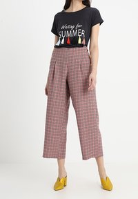 Černé tričko s nápisem "Čekání na LÉTO" a barevnými třásněmi, které je kombinováno s vysokým pasem, širokými kalhotami v červeno-černé kostce a žlutými vysokými podpatky.