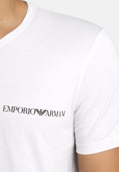 Κοντινό πλάνο ενός ατόμου που φοράει λευκό μπλουζάκι με μικρό μαύρο λογότυπο Emporio Armani τυπωμένο στο στήθος.