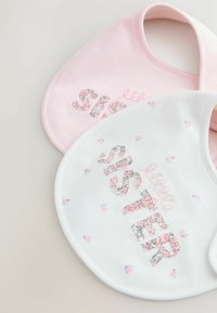Două bavete pentru bebeluși din bumbac: una roz cu inscripția „surioară” pe un material floral, cealaltă albă cu cuvântul „soră” și modele florale. Design rotunjit.