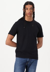 Jeune homme avec de longues tresses portant un t-shirt noir uni et un jean bleu, debout les mains derrière le dos sur un fond blanc.