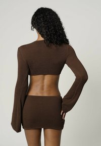 Femme aux cheveux bouclés et foncés vue de dos, portant un crop top en maille marron à manches longues et une jupe mini assortie, sur fond uni.