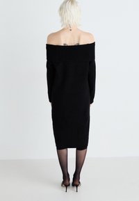 Vestito a maglia nero off-the-shoulder con maniche lunghe, silhouette aderente e lunghezza al ginocchio. Indossato con collant neri trasparenti e scarpe con tacco alto.