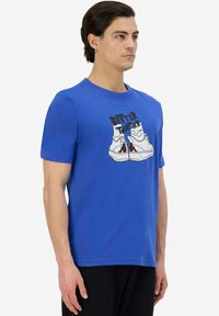 T-shirt in cotone blu con collo a giro, caratterizzata da un design grafico di scarpe e il testo "BETTER TOGETHER" in nero e rosso.