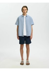 Jeune homme portant une chemise à manches courtes rayée sur un t-shirt blanc, un short bleu marine et des sandales beiges, sur un fond clair uni.