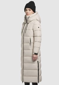 Lange beige pufferjas met ritssluiting aan de voorkant, hoge kraag en trekkoorden. Heeft een horizontaal gestikt ontwerp, zijrits en zwarte accenten.