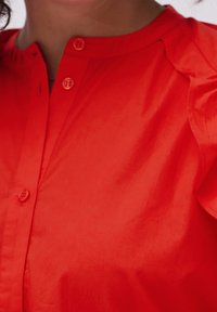 Camicia rossa con colletto rotondo, caratterizzata da bottoni sul davanti. Realizzata in tessuto leggero, con una trama liscia e una leggera lucentezza.