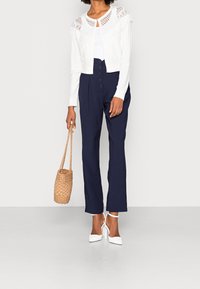 Bílý krátký kardigan s krajkovými detaily, v kombinaci s tmavě modrými vysokými kalhotami a bílými sandály na podpatku. Doplňky: pletená kabelka.