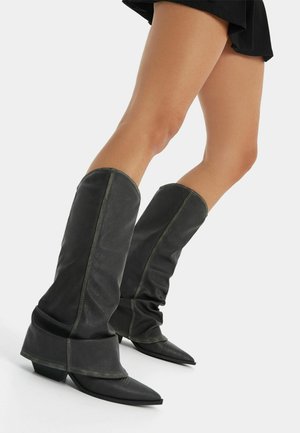 Persona che indossa stivali di pelle neri morbidi al ginocchio, con punte appuntite e tacchi bassi, abbinati a una gonna nera su sfondo bianco.