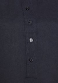 Gros plan d'une chemise en tissu sombre avec quatre boutons noirs cousus sur la patte centrale.