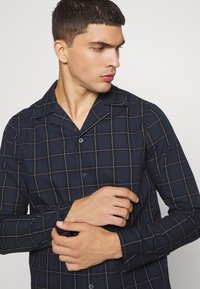 Camisa de manga larga de color azul marino con un patrón a cuadros en blanco. Presenta un cuello, un cierre frontal con botones y una textura sutil.