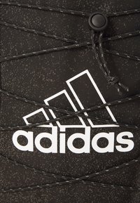 Juodas sportinis batelis su tekstūruotu viršumi, su baltu Adidas logotipu ir juodomis elastinėmis raištelėmis. Matomas taškuotas raštas medžiagoje.