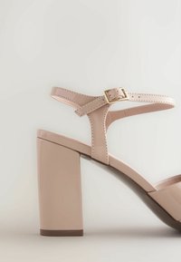 Sandale à talon haut en cuir verni nude avec talon carré et bride de cheville réglable avec boucle dorée sur fond neutre.
