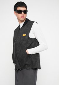 Vestă neagră cu model, cu fermoar frontal și două buzunare laterale. Are un guler negru simplu și o etichetă galbenă pe piept.