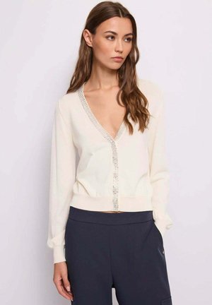 Giovane donna con lunghi capelli castani che indossa un cardigan bianco a scollo a V con strass e pantaloni scuri a vita alta, che guarda a sinistra.