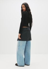 Schwarzes, strukturiertes Langarmoberteil, kombiniert mit einem schwarzen Wickelrock und locker sitzenden, hellblauen weitem Jeans. Das Model steht mit dem Rücken zur Kamera vor einem weißen Hintergrund.