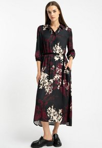 Kobieta w czarno-burgundowej sukience koszulowej midi w kwiaty z paskiem, w połączeniu z czarnymi mokasynami, stojąca na białym tle.
