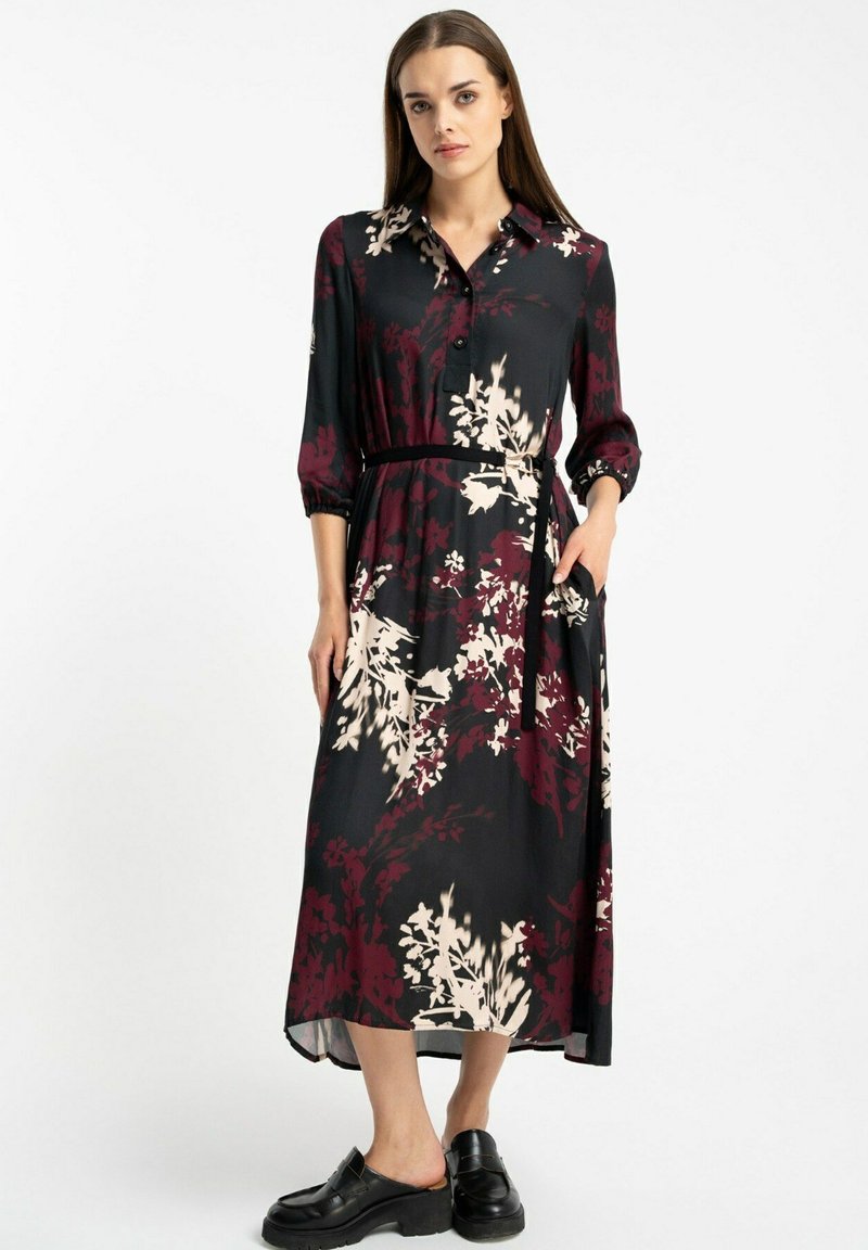 Kobieta w czarno-burgundowej sukience koszulowej midi w kwiaty z paskiem, w połączeniu z czarnymi mokasynami, stojąca na białym tle.
