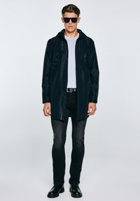 Jauns vīrietis, valkājot melnu kapuci ar rāvējslēdzēju, pelēku krekla, melnas džinsu bikses, melnu jostu, melnus zābakus un melnus saulesbrilles, stāv pret vienveidīgu fonu.