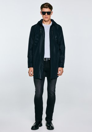 Młody mężczyzna w czarnej kurtce z kapturem, szarej koszuli, czarnych dżinsach, czarnym pasku, czarnych butach i czarnych okularach przeciwsłonecznych stoi na tle jednolitego tła.