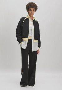 Zwarte jas met gele accenten, gecombineerd met een witte overhemden en zwarte wijde broek, met een gladde textuur en casual ontwerp.