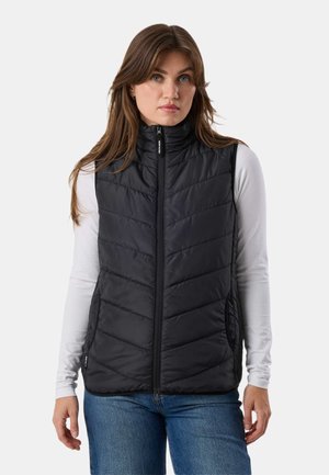 Kobieta o brązowych włosach, ubrana w czarną pikowaną kamizelkę na białej koszuli z długim rękawem i niebieskie jeansy, stojąca na jednolitym tle.