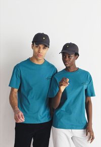 T-shirts en coton teal à manches courtes, ornés d'un petit logo. Casquettes noires avec un emblème jaune, portées par deux silhouettes sur un fond uni.