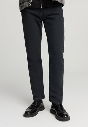 Pantaloni in denim nero con vestibilità slim e classico design a cinque tasche, abbinati a eleganti scarpe in pelle nera. Texture liscia, dettagli minimi.