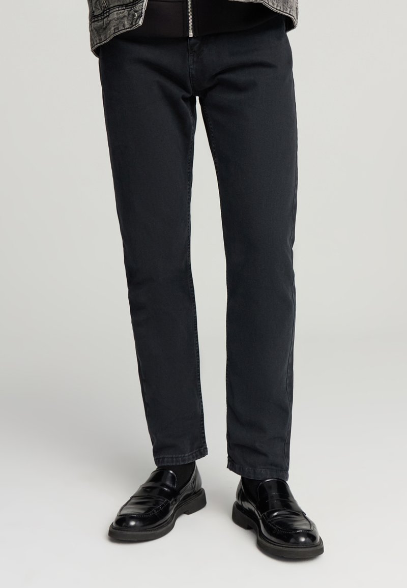 Pantaloni in denim nero con vestibilità slim e classico design a cinque tasche, abbinati a eleganti scarpe in pelle nera. Texture liscia, dettagli minimi.