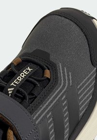 Chaussure de sport noire avec tige en maille, détails texturés et trois rayures diagonales sombres. Présente une boucle de lacet marron et des accents de semelle blanche.